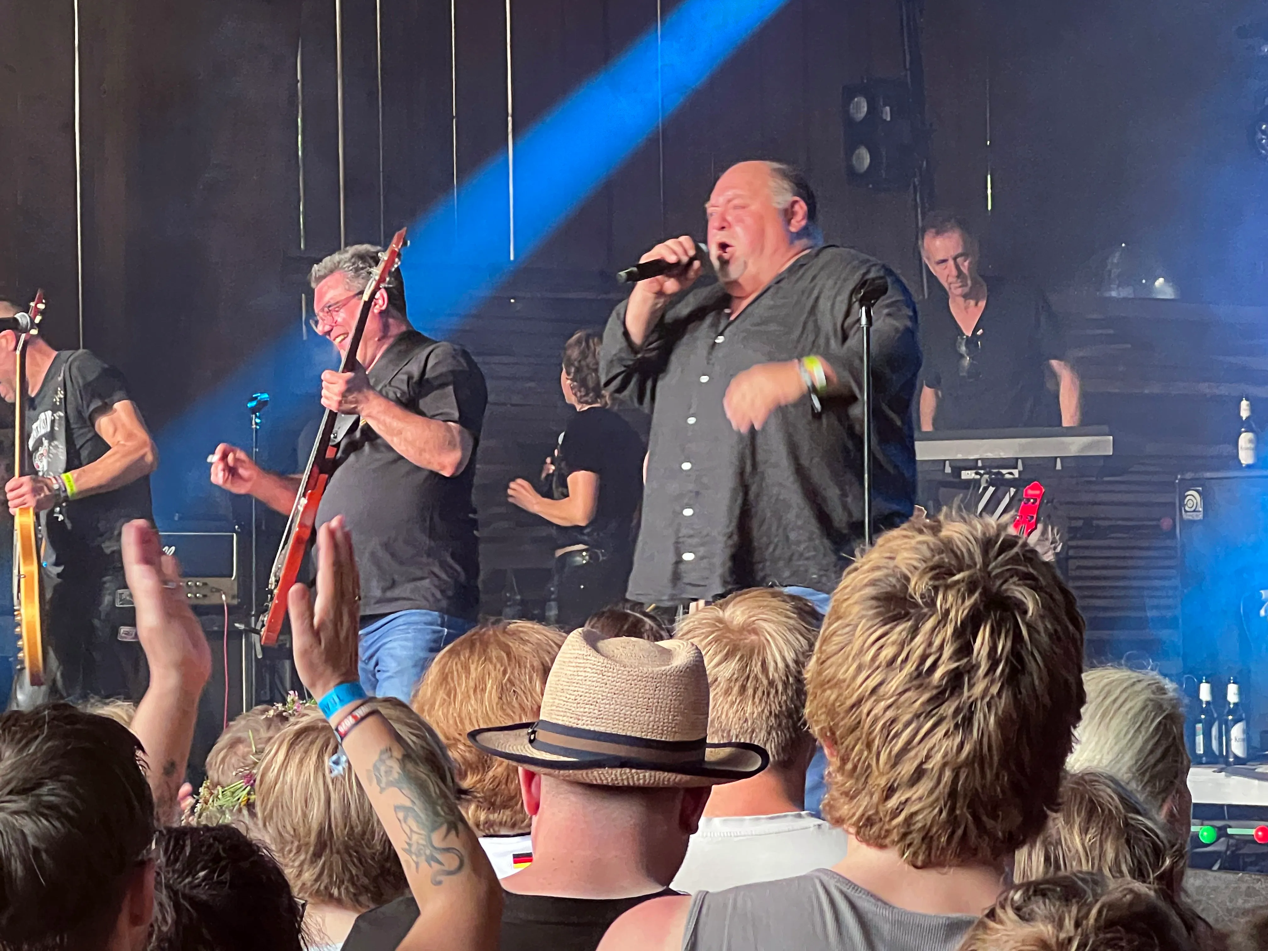 Bühnenbild des I-Rock Festivals mit einer Band während der Vorstellung. Der Sänger mit Mikrofon steht im Vordergrund, während die Bandmitglieder im Hintergrund spielen und das Publikum jubelt.