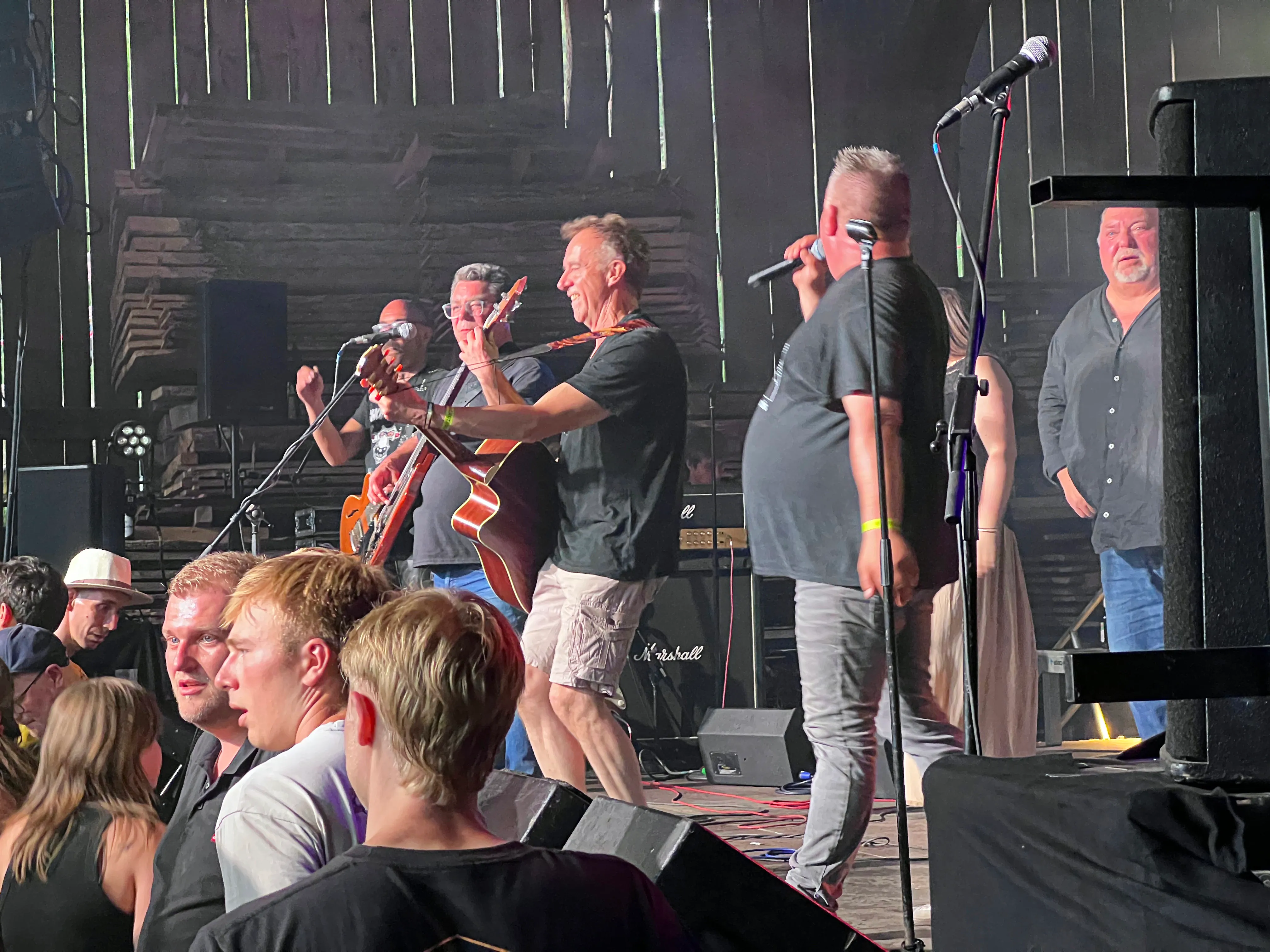 Band spielt auf der Bühne des I-Rock Festivals in Plettenberg. Ein Mann singt in ein Mikrofon, während ein anderer Gitarre spielt, umgeben von weiteren Bandmitgliedern und Technik.