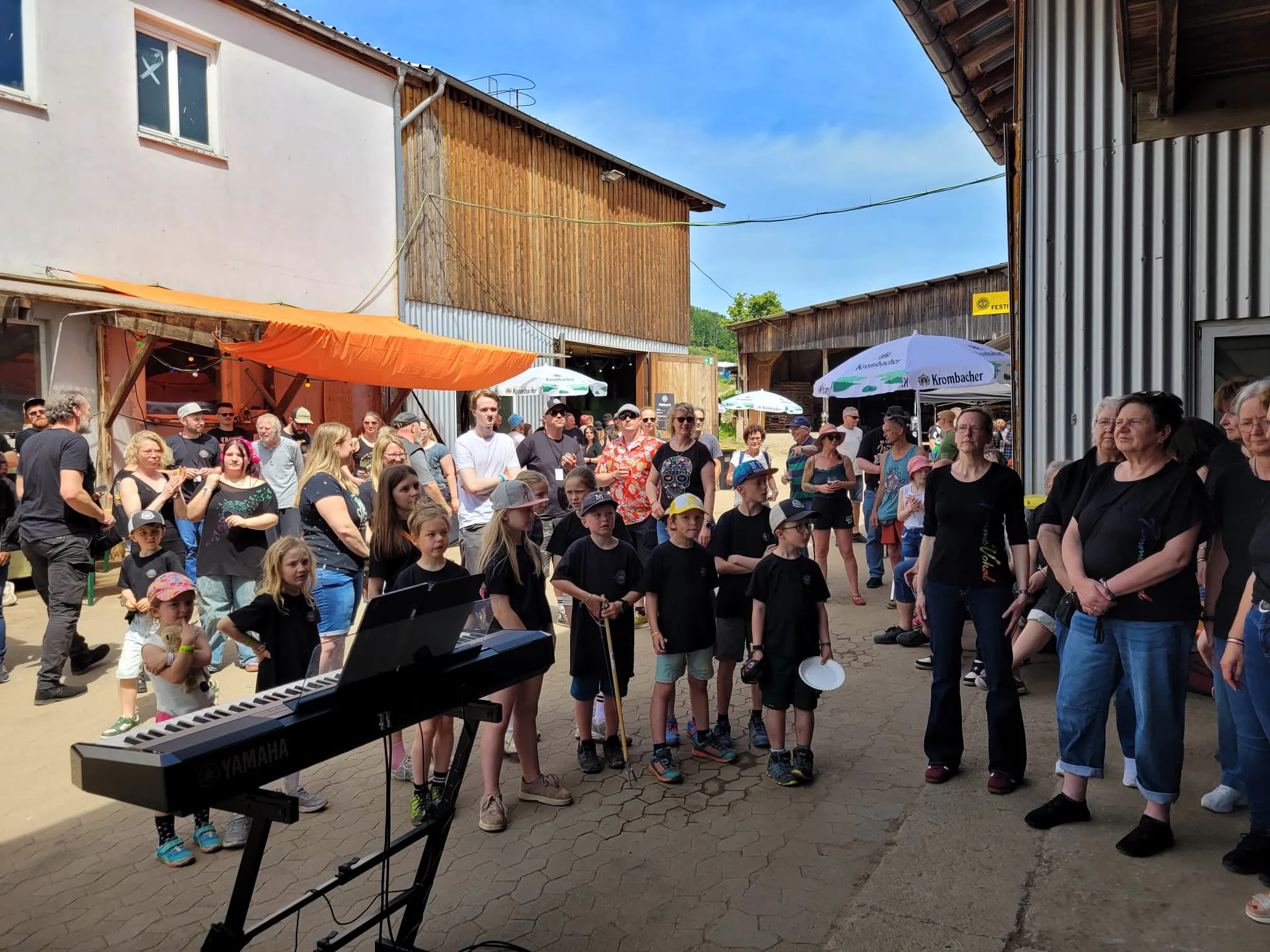 Eine Gruppe von Kindern und Jugendlichen spielt vor einem Publikum auf einem Festivalgelände mit ländlichem Hintergrund auf Instrumenten wie Keyboards und Trompeten.
