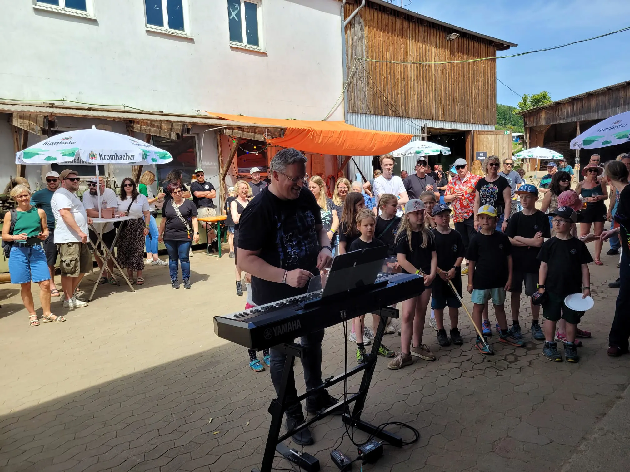 Ein Musiker spielt Keyboard vor einer Gruppe von Kindern und Erwachsenen im Freien. Im Hintergrund sind Gebäude und Festivalzelte zu sehen.