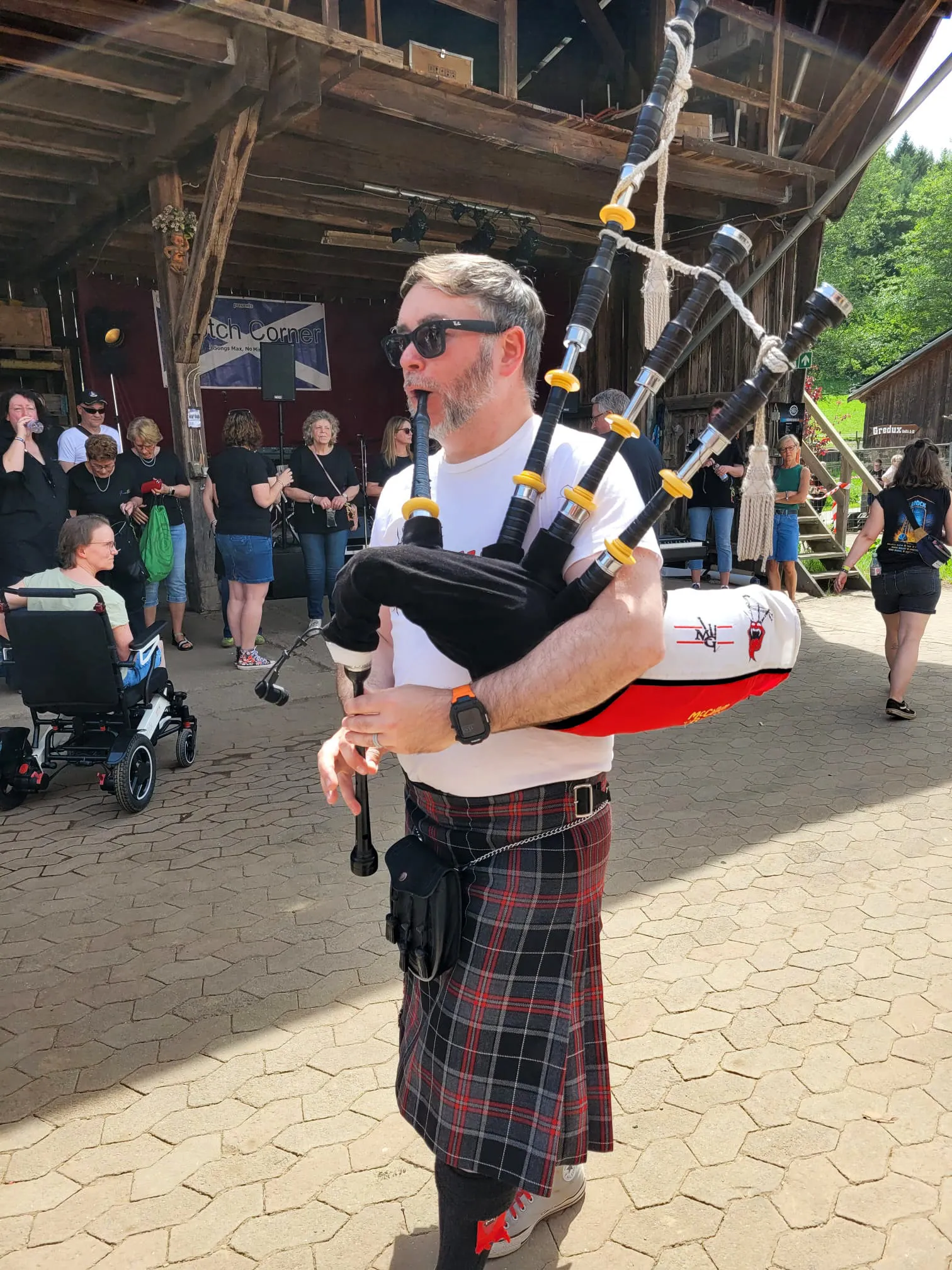 Ein Dudelsackspieler in traditioneller schottischer Kleidung spielt vor einer Bühne im Freien. Im Hintergrund stehen und sitzen weitere Festivalbesucher.
