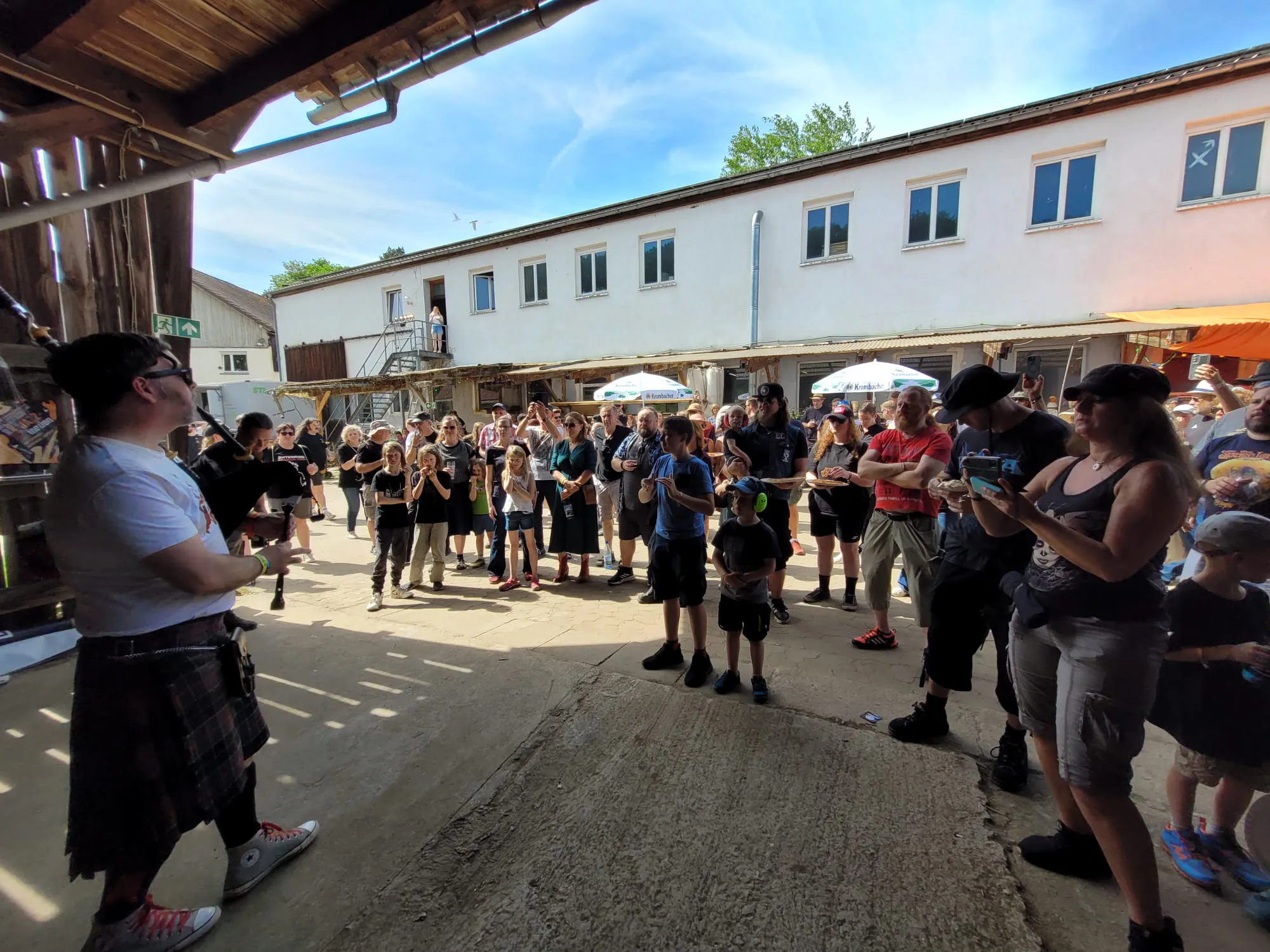 Eine Gruppe von Menschen steht vor einem Gebäude mit mehreren Fenstern und beobachtet einen Mann in einem Kilt, der eine Dudelsack spielt.