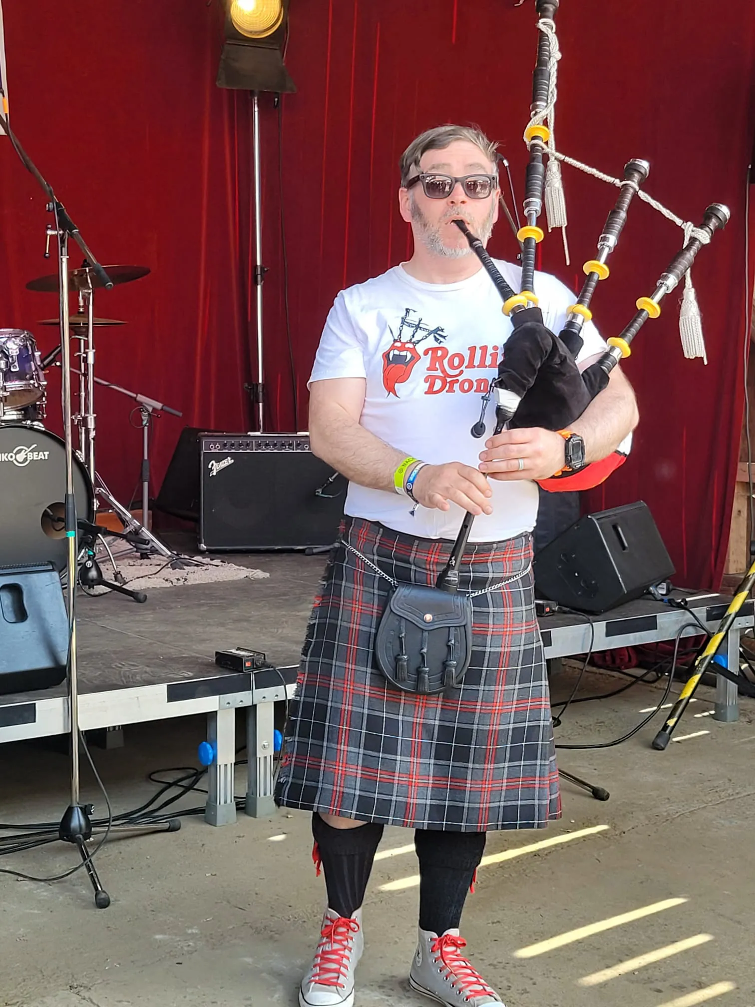 Ein Mann mit Bart spielt auf einer Dudelsack auf einer Bühne vor rotem Hintergrund. Er trägt einen Kilt, Socken und Turnschuhe.