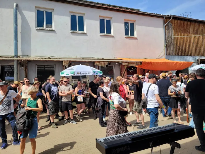 Menschen stehen und sitzen vor einer Bühne im Freien, einige tanzen vor einem Keyboard. Im Hintergrund sind Gebäude und Werbeschilder zu sehen.