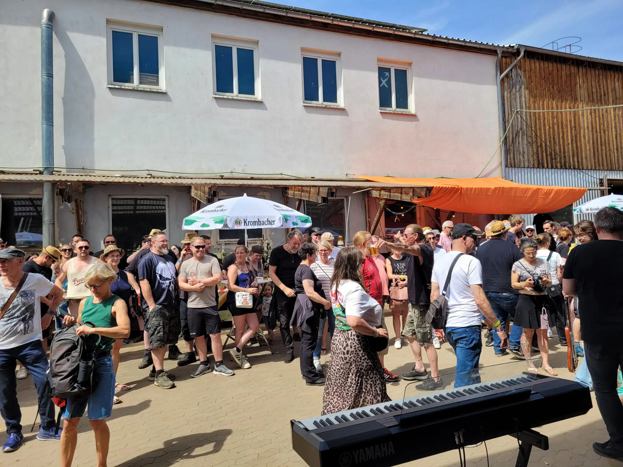 Menschen stehen und sitzen vor einer Bühne im Freien, einige tanzen vor einem Keyboard. Im Hintergrund sind Gebäude und Werbeschilder zu sehen.