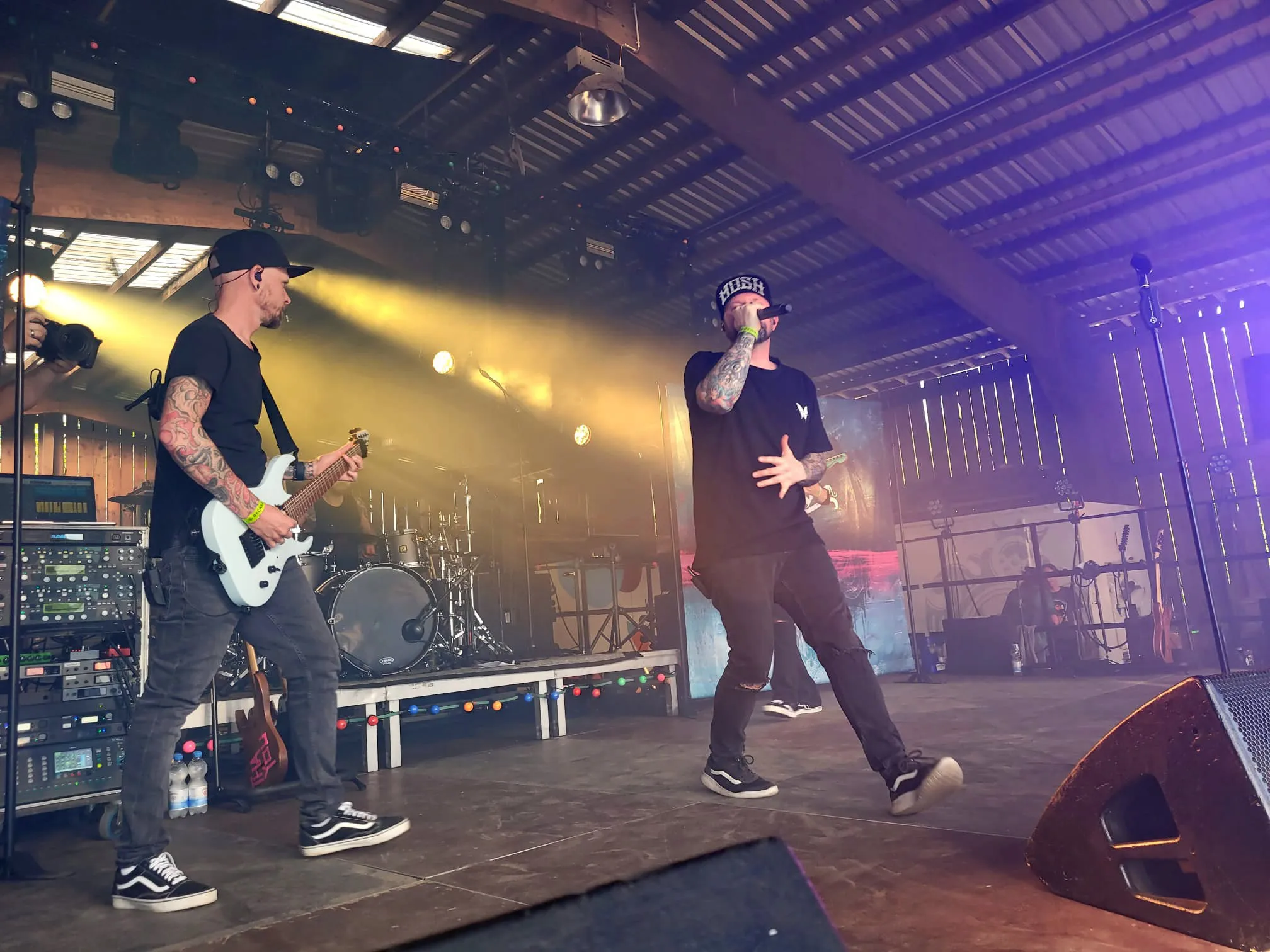 Zwei Musiker auf der Bühne des I-Rock Festivals in Plettenberg. Links spielt ein Mann Gitarre, rechts steht ein Mann mit Mütze und Mikrofon vor den Zuschauern.