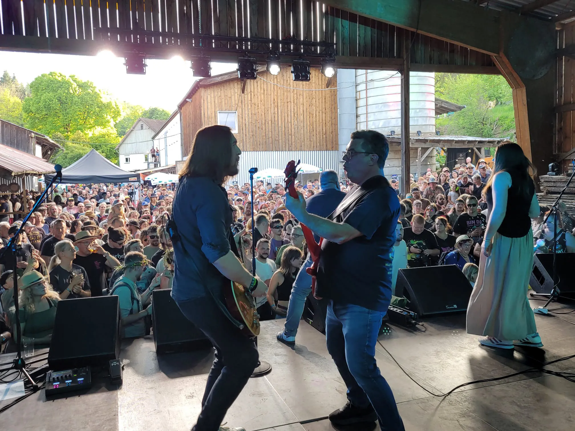 Eine Band spielt vor einer großen Menschenmenge auf der Bühne eines Open-Air-Festivals. Im Hintergrund ein Holzgebäude und Bäume.