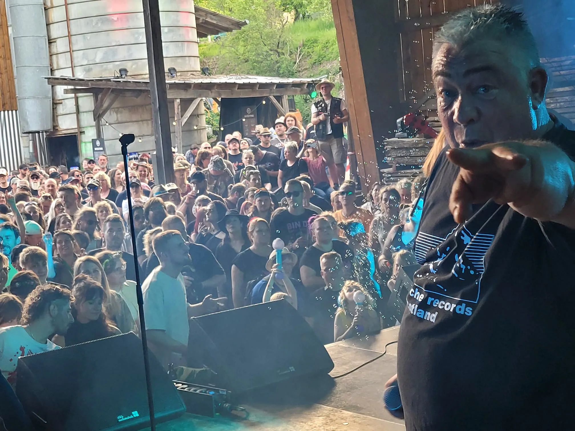 Ein Mann bläst Seifenblasen vor eine große Menschenmenge auf einem Open-Air-Festival. Im Hintergrund eine Bühne und Gebäude.