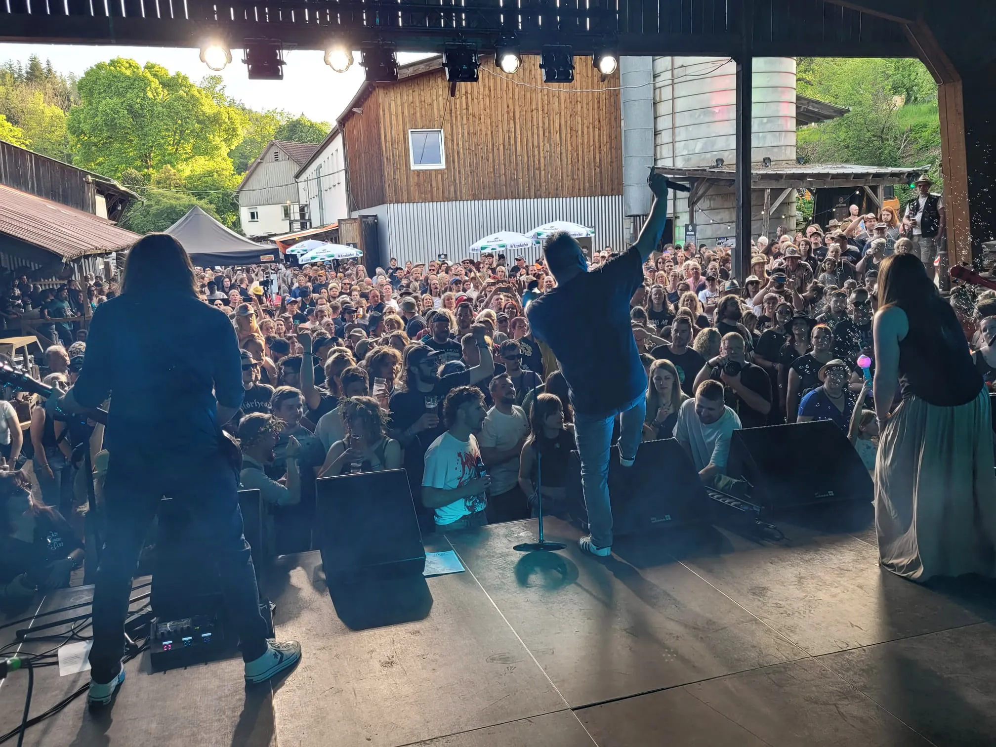 Blick über ein dichtes Publikum bei einem Open-Air-Konzert. Auf der Bühne steht ein Musiker mit Mikrofon und eine Frau, während im Hintergrund ein Holzgebäude und Zelte zu sehen sind.