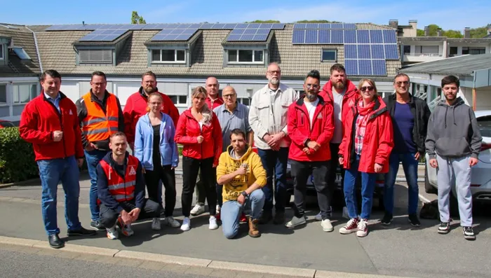Eine Gruppe von Menschen, viele in roten Jacken, steht vor einem Gebäude mit Solarpaneelen auf dem Dach.