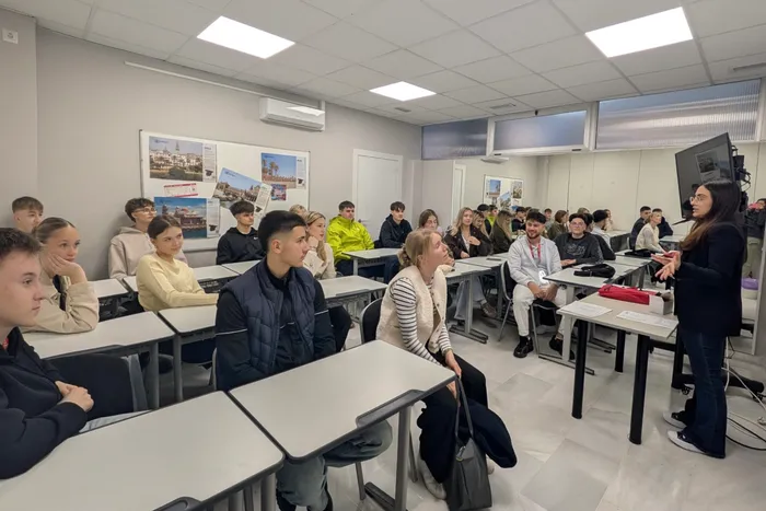 Schülergruppe in einem hellen Klassenraum lauscht einer Vortragenden. Im Hintergrund sind Poster mit Bildern von Málaga zu sehen.