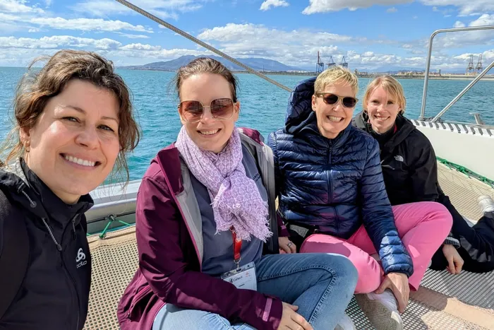 Vier lächelnde Frauen sitzen an Deck eines Bootes vor Küstenlandschaft und Bergen. Sie absolvieren ein berufliches Praktikum in Málaga.