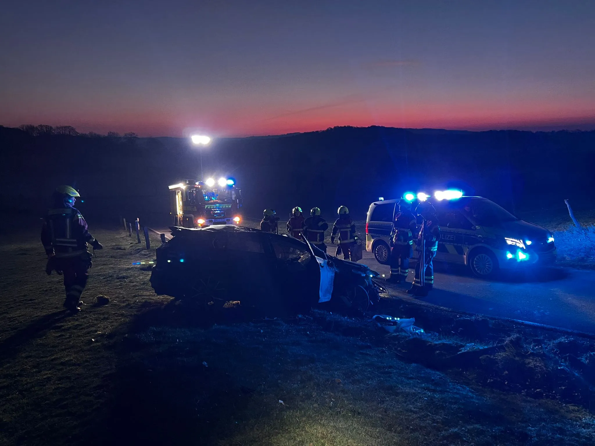 Polizei- und Feuerwehrleute sichern Unfallstelle mit einem stark beschädigten Auto in der Dämmerung.