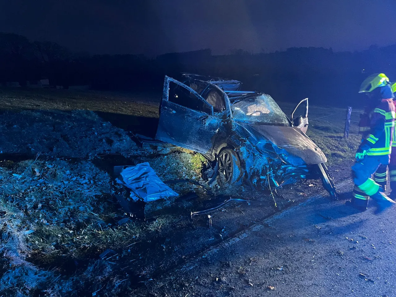 Ein stark beschädigtes Auto nach einem Unfall am Straßenrand. Ein Feuerwehrmann steht daneben und betrachtet die Schäden.