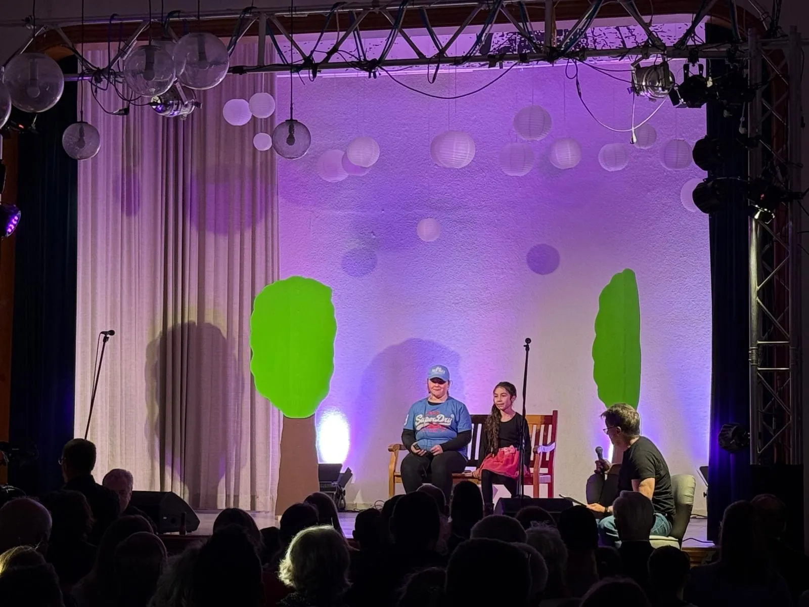 Drei Kinder sitzen auf Stühlen auf einer Bühne vor einem Publikum. Im Hintergrund eine Kulisse mit grünen Blättern und weißen Papierlaternen.