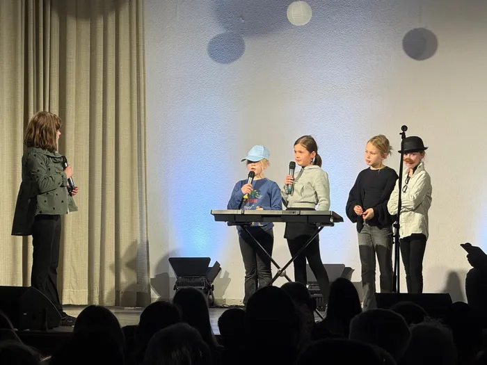 Vier Kinder stehen auf einer Bühne vor einem Publikum und singen in Mikrofone. Eine Frau steht seitlich und spricht ebenfalls in ein Mikrofon.