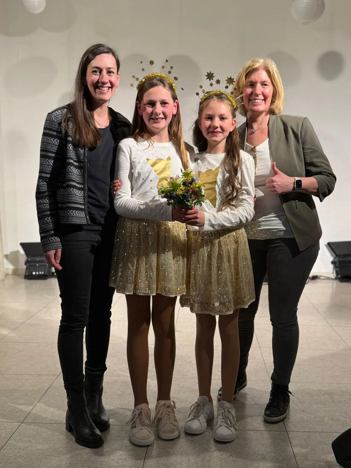 Zwei Mädchen mit Sternenkrönchen und Blumenstrauß stehen zwischen zwei erwachsenen Frauen auf einer Bühne. Im Hintergrund ist ein helles Bühnenbild zu sehen.
