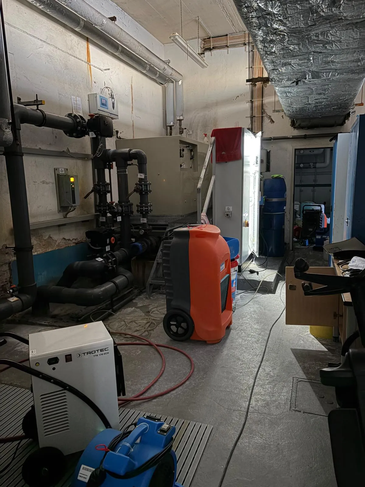 Technikraum einer Schwimmhalle mit Rohren, Pumpen und Geräten zur Wasseraufbereitung. Im Vordergrund ein Luftentfeuchter der Marke Trotec.