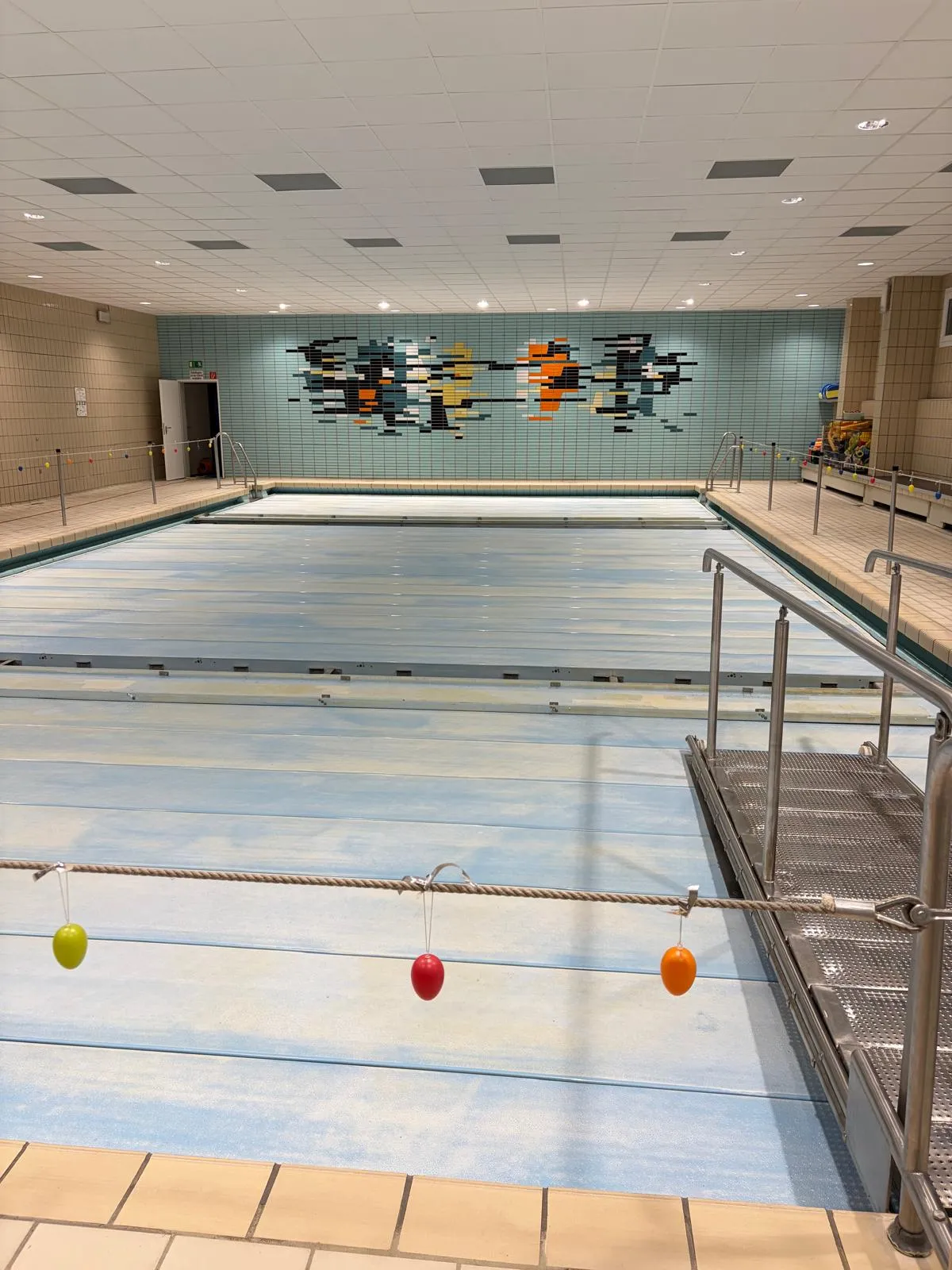 Leeres Schwimmbecken mit einer abstrakten Wandgestaltung im Hintergrund. Eine Rampe führt ins Wasser, drei Bälle hängen an einer Leine.