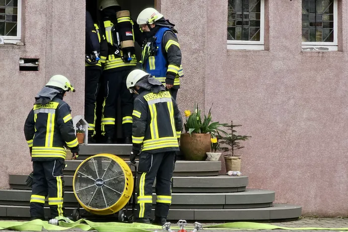 Mehrere Feuerwehrleute in voller Montur stehen vor einem Gebäude, während ein Ventilator Rauch abführt. Schläuche liegen auf dem Boden.