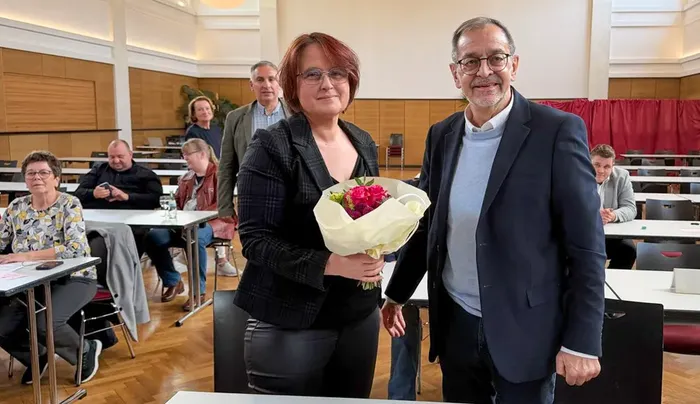 Eva Kitz hält einen Blumenstrauß entgegen, während Michael Scheffler neben ihr steht. Im Hintergrund sind Tische und Stühle in einem Veranstaltungsraum zu sehen.