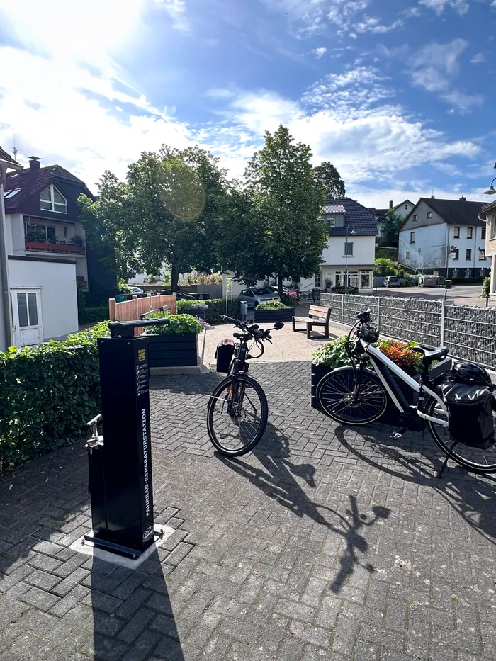 Dorfplatz mit Fahrradständer und zwei Fahrrädern, im Hintergrund Häuser und Bäume an einem sonnigen Tag.