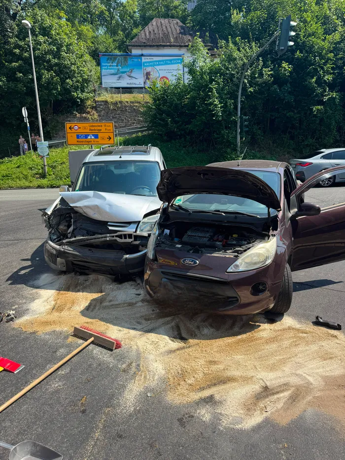 Zwei stark beschädigte Autos blockieren eine Kreuzung nach einem Unfall. Im Hintergrund sind weitere Fahrzeuge und Verkehrsschilder zu sehen.