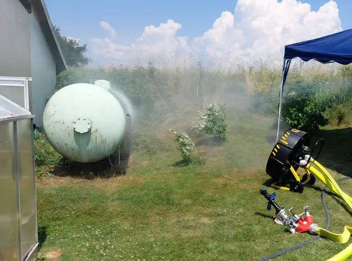 Dampf steigt von einem großen, hellgrünen Tank auf einer Wiese auf. Im Vordergrund steht eine Feuerwehrausrüstung mit Schläuchen unter einem blauen Pavillon.