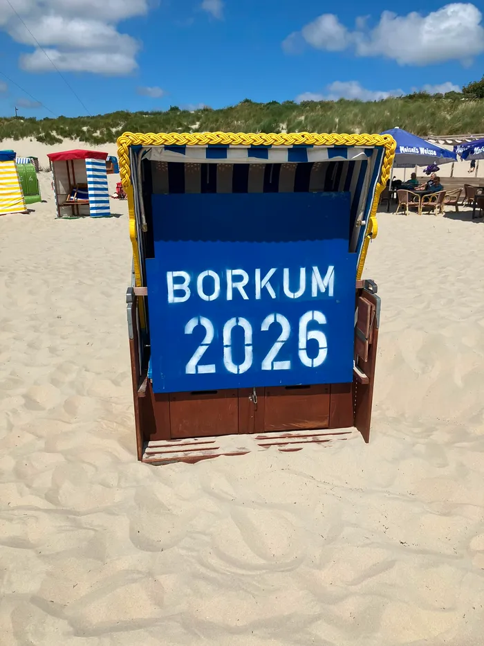 Ein blauer Strandkorb mit der weißen Aufschrift „BORKUM 2026“ steht auf einem Sandstrand vor Dünen.