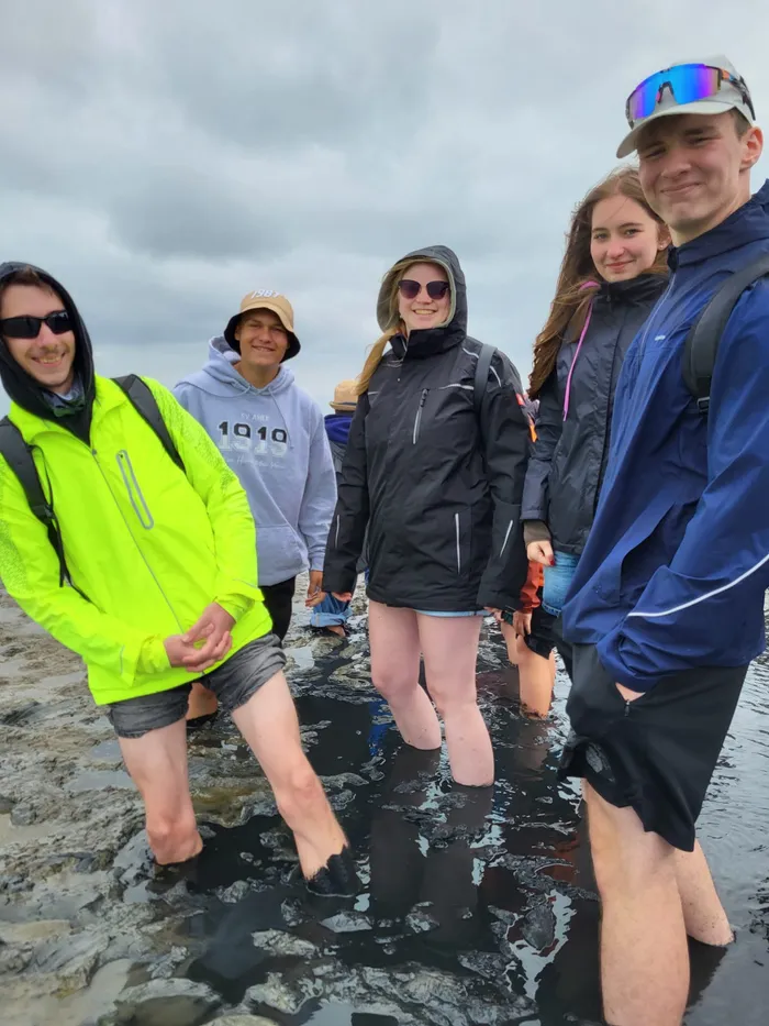 Eine Gruppe von Menschen in Outdoor-Kleidung steht in flachem Wasser an einem felsigen Ufer unter bewölktem Himmel.