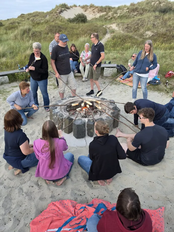 Eine Gruppe von Menschen sitzt und steht im Kreis um eine Feuerstelle im Sand und röstet am Stock über dem Feuer.