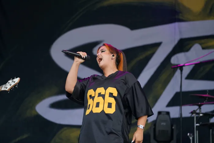 Sängerin mit orangefarbenen Haarsträhnen singt in ein Mikrofon und trägt ein schwarzes Trikot mit der gelben Nummer 666.