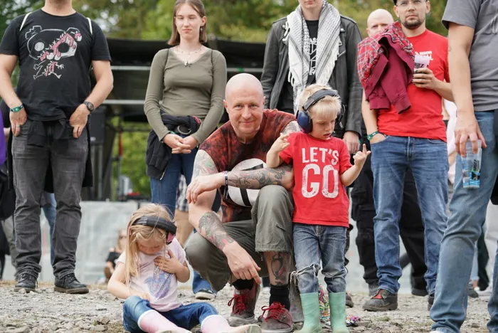 Ein tätowierter Mann hockt mit zwei Kindern, die Kopfhörer tragen, vor einer Menschenmenge im Freien.