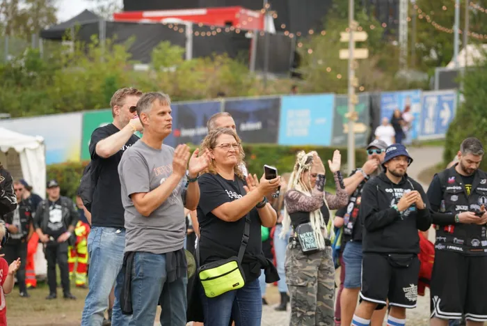 Menschen applaudieren bei einem Open-Air-Event vor bunten Bannern und Zelten.