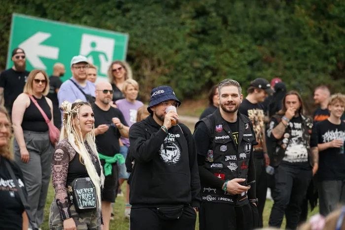 Menschenmenge bei einem Event, im Vordergrund ein Mann in einem schwarzen Kapuzenpullover beim Trinken und eine lächelnde Frau mit blonden Dreadlocks.
