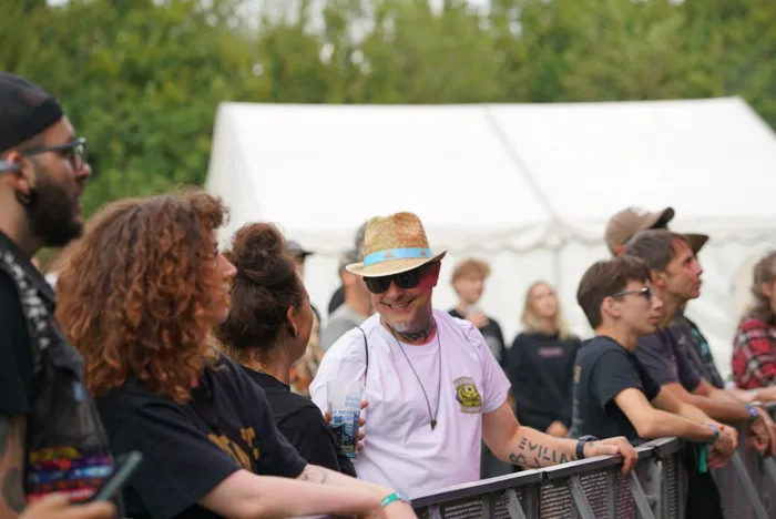 Ein lächelnder Mann mit Strohhut und Sonnenbrille steht hinter einer Metallabsperrung vor einem weißen Zelt bei einer Veranstaltung.