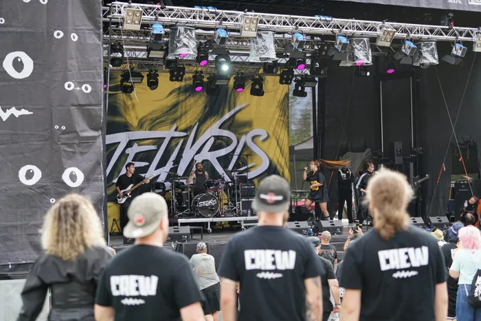 Die Band Tellvris spielt auf einer Bühne, während im Vordergrund Crew-Mitglieder mit „CREW“-Aufdruck auf dem Rücken stehen.