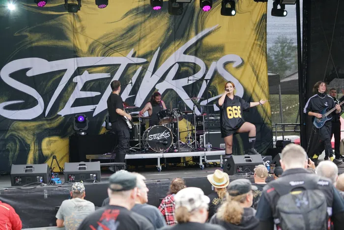 Die Band Stellvris tritt live auf einer Bühne vor einem gelb-schwarzen Backdrop auf. Eine Sängerin in einem schwarzen Shirt mit der Aufschrift „666“ singt.