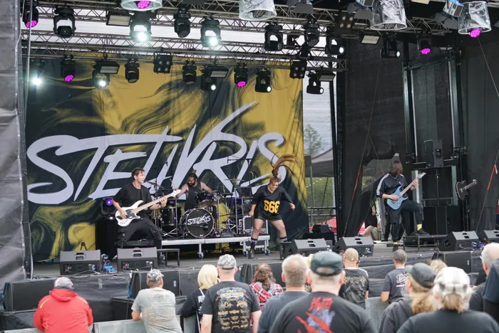Die Band Stellvris tritt auf einer Bühne vor einem großen Banner mit ihrem Namen auf. Musiker spielen Gitarre und Schlagzeug vor einem Publikum.
