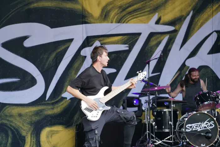 Ein Musiker spielt eine weiße Bassgitarre vor einem großflächigen Stellvris-Banner. Im Hintergrund ist ein Schlagzeuger zu sehen.