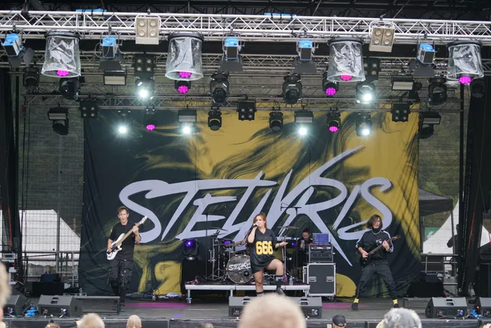 Die Band Stellvris tritt auf einer Bühne vor einem großflächigen gelb-schwarzen Backdrop auf. Im Zentrum singt eine Frau, flankiert von zwei Gitarristen.