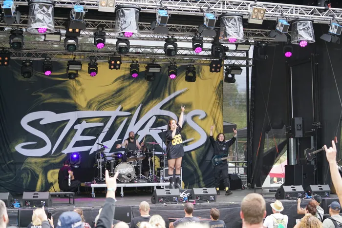 Die Band STEHKRS tritt auf einer Bühne vor einem großen gelb-schwarzen Backdrop auf, während die Sängerin die Arme hebt.