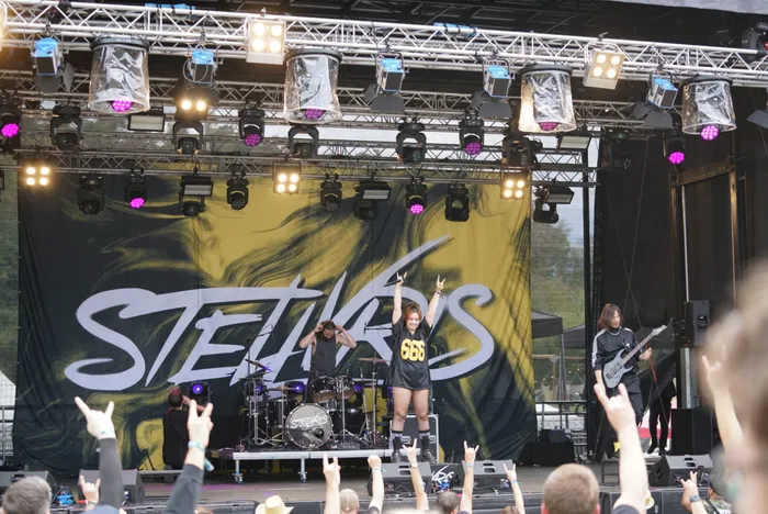 Die Band Steelwris tritt auf einer Bühne vor einem großen Banner auf. Die Sängerin steht im Zentrum und macht Handzeichen.
