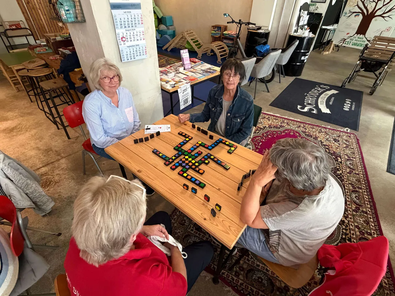 Vier Personen sitzen an einem Holztisch und spielen ein Brettspiel mit bunten Steinen.