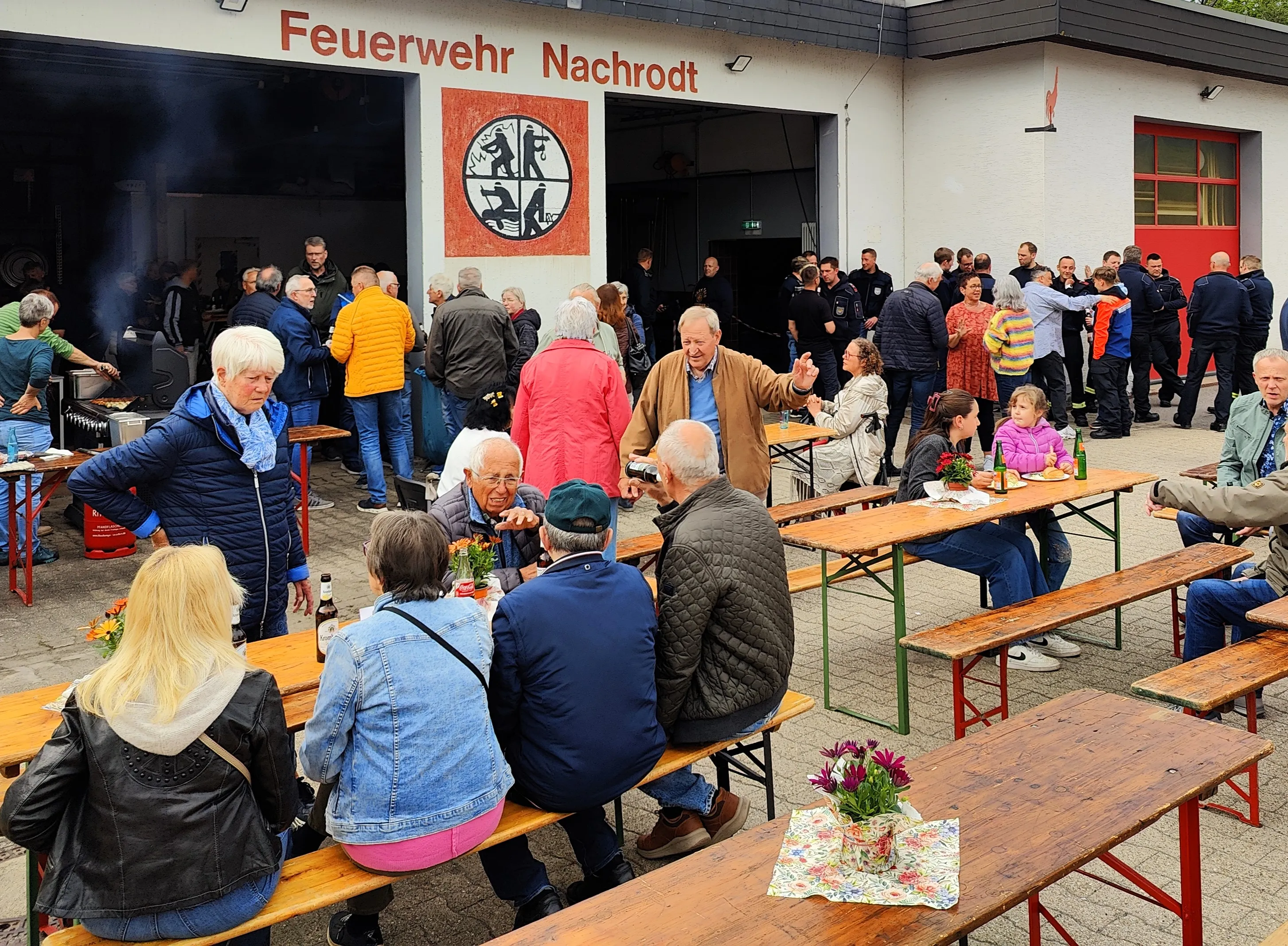 Menschen sitzen und stehen vor dem Gebäude der Freiwilligen Feuerwehr Nachrodt-Wiblingwerde zusammen, einige grillen und essen an Biertischgarnituren im Freien.
