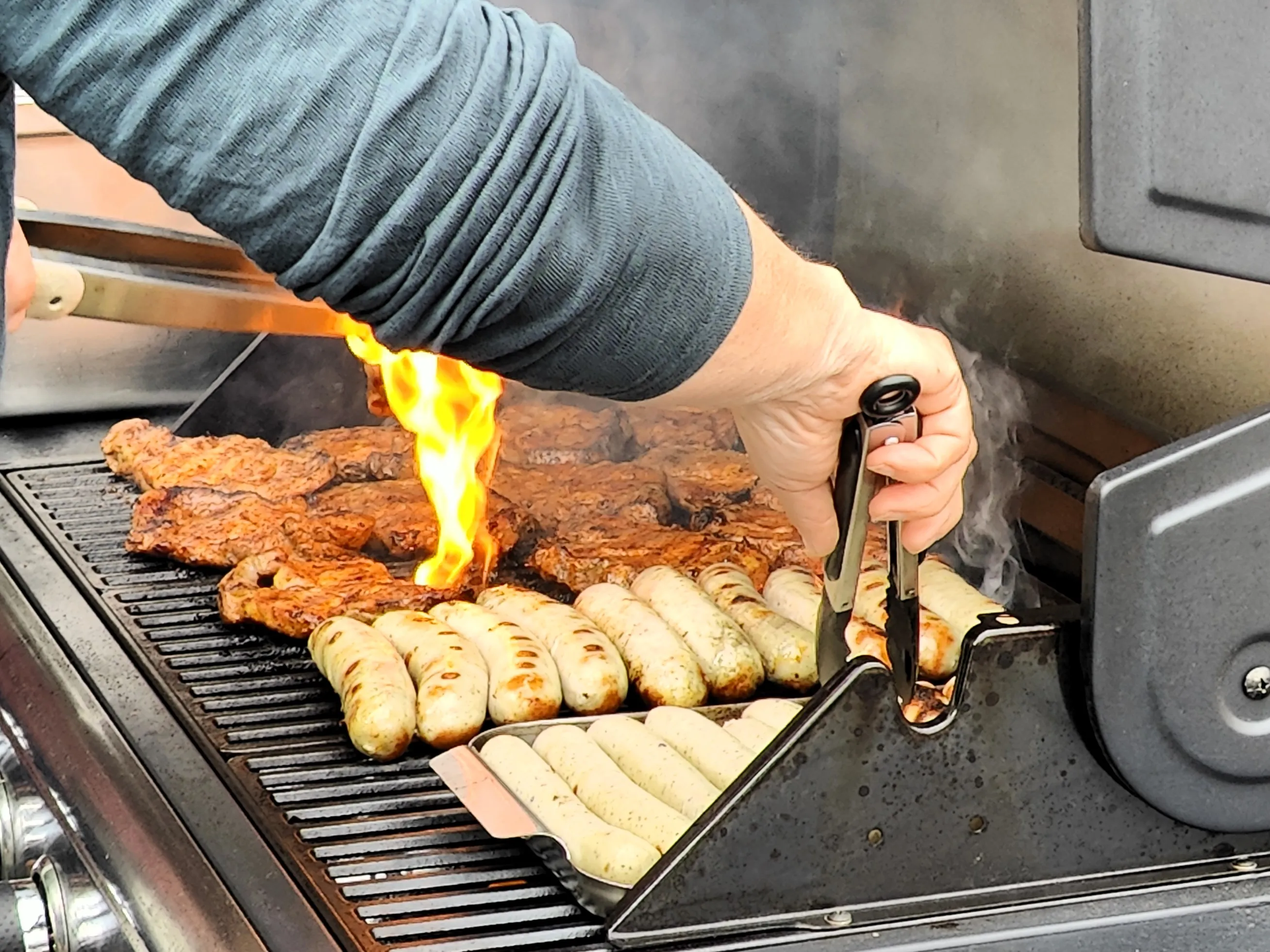 Grill mit Fleisch und Würstchen, während eine Person mit einer Zange Lebensmittel auf dem Grill platziert. Im Hintergrund ist Rauch zu sehen.