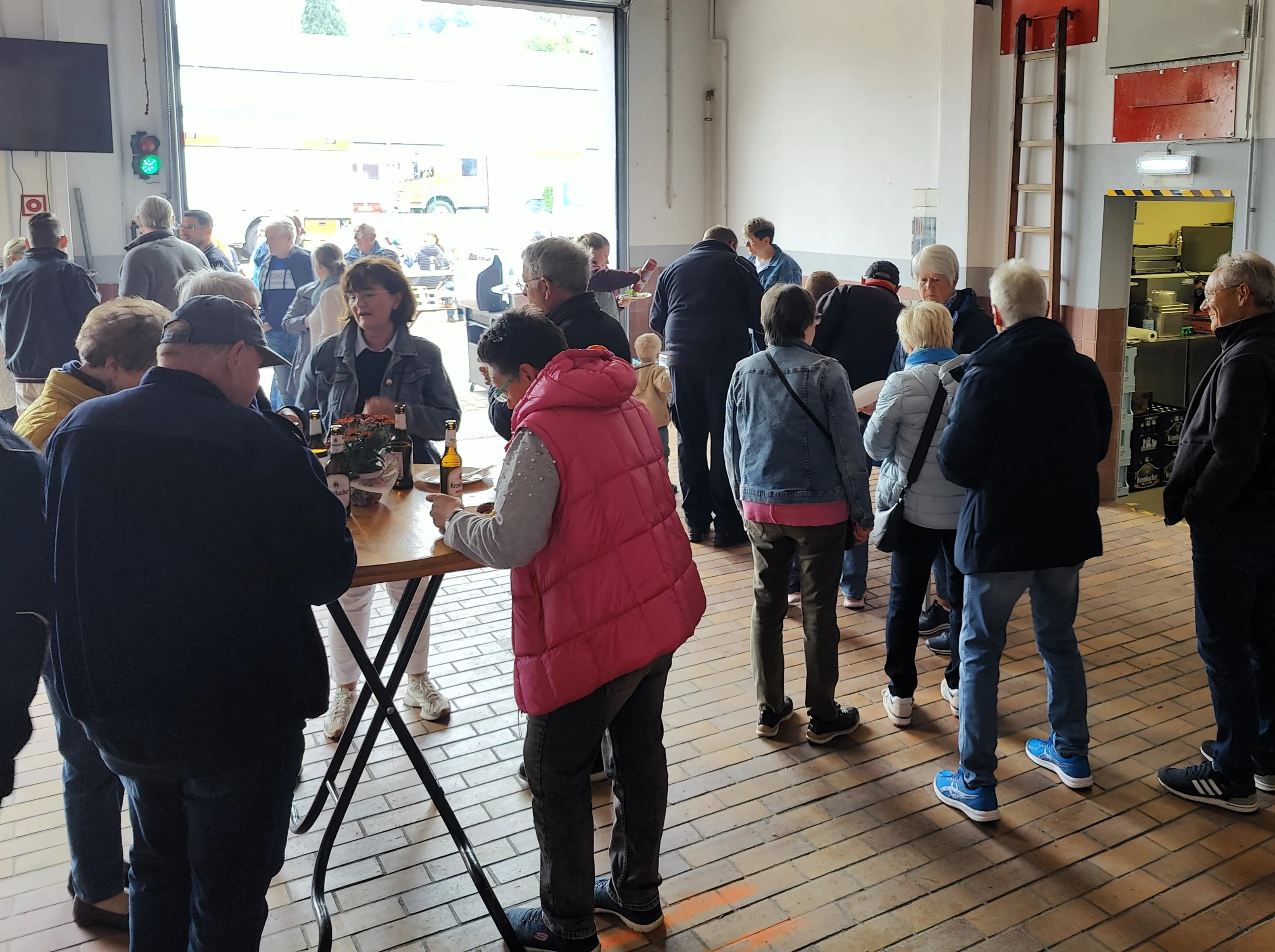 Menschen stehen und unterhalten sich bei einem Grillfest in einer großen Halle mit offenen Türen.