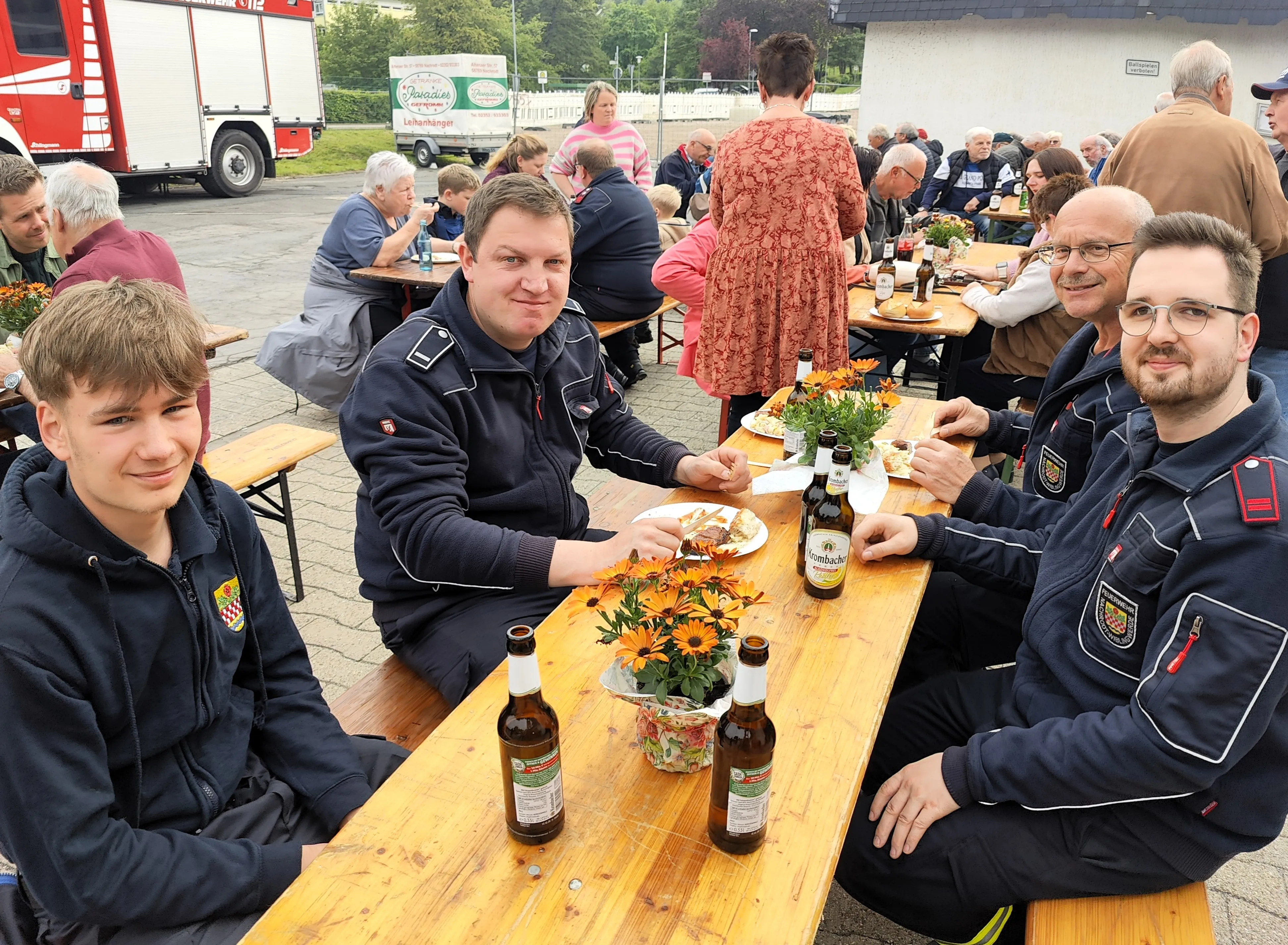 Mehrere Feuerwehrleute und andere Gäste sitzen an Biertischgarnituren und essen im Freien. Im Hintergrund steht ein rotes Feuerwehrauto.