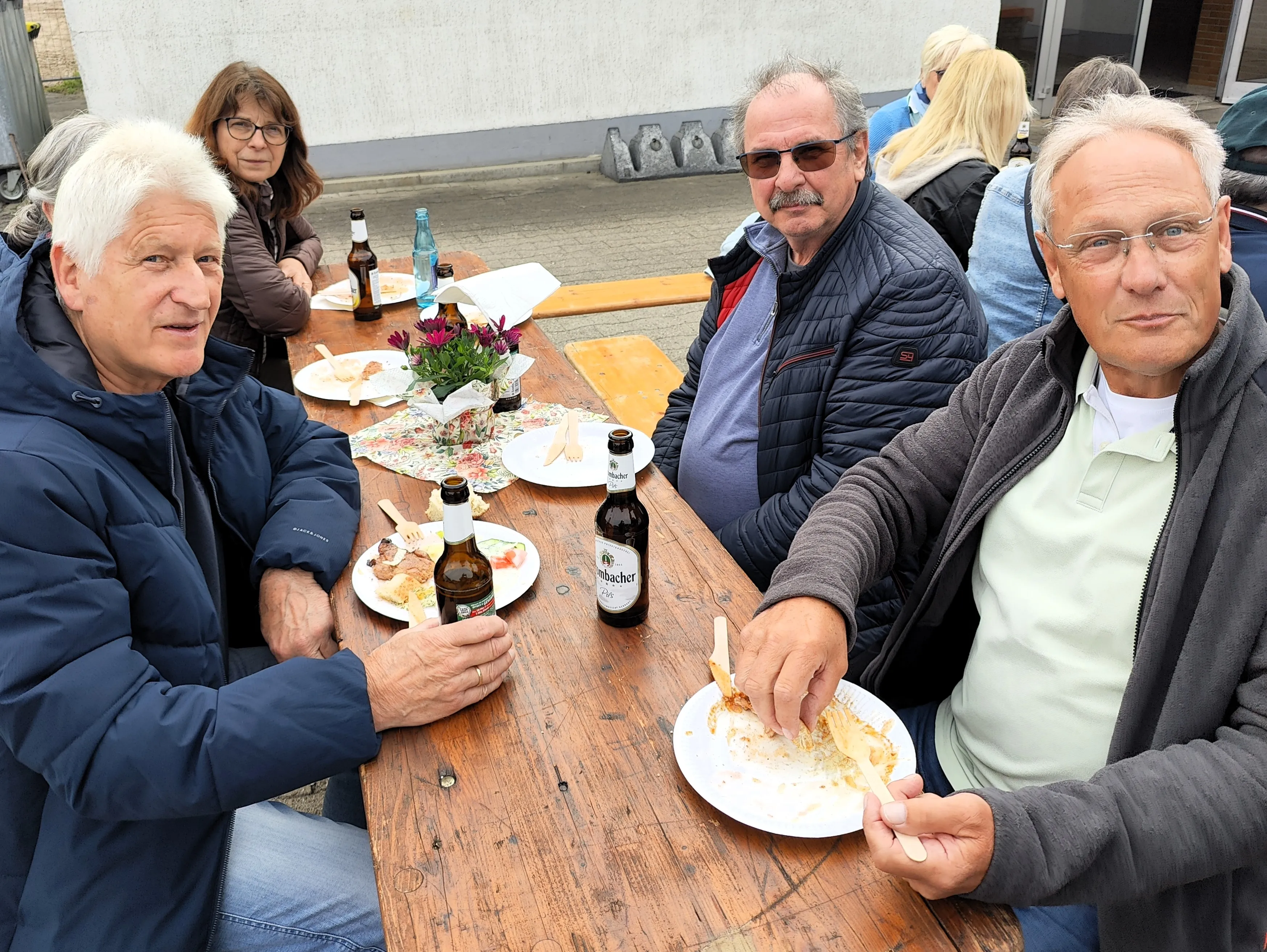 Mehrere ältere Menschen sitzen an einem Holztisch im Freien und essen. Auf dem Tisch stehen Getränke und Essensreste.
