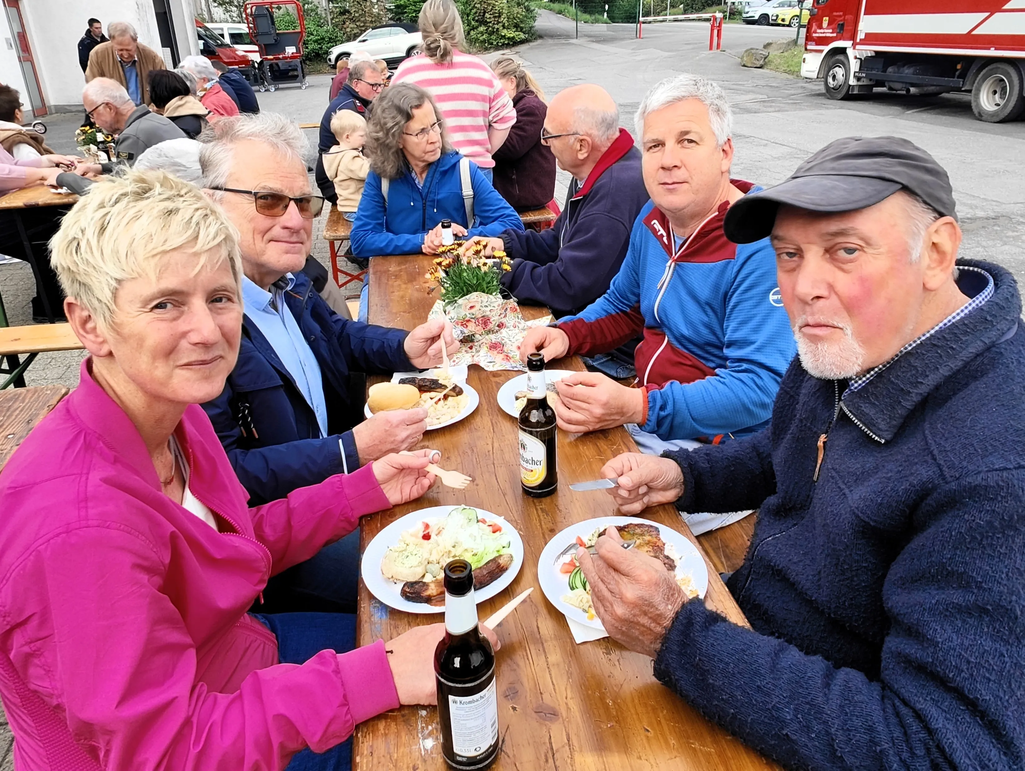 Gruppe von Menschen sitzt an einem Holztisch und isst. Auf dem Tisch stehen Getränke und Salate, im Hintergrund ein rotes Feuerwehrauto.